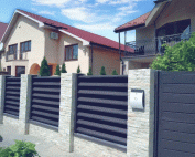 Fence Blinds Premium 4 Modern residential area with a stylish dark gray metal fence and stone pillars, red-roofed houses, and greenery under a clear sky.